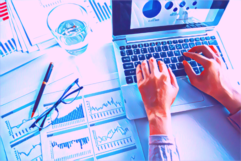 Close up of hands typing a report on a laptop. Next to the laptop are pieces of paper showing charts, a glass of water, a pen and a pair of glasses